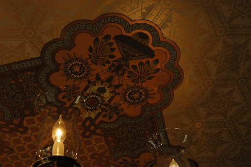 An ornate ceiling design featuring floral patterns and geometric shapes with intricate detailing. The central focus is a vivid floral motif surrounded by layers of patterns in earthy tones. Hanging below is a lit glass lamp with a warm glow, casting subtle shadows and highlighting the ornate ceiling.