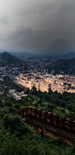 A large historical fort complex is set amidst a lush, green landscape with rolling hills. The fort's architectural structures are prominently featured, displaying intricate designs and an expansive layout. The sky above is overcast, suggesting a moody or impending weather change.