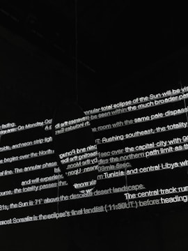 Bright white text is projected onto a dark surface, with several lines overlapping in various orientations. The text appears fragmented and partially obscured by shadows, creating an abstract and mysterious visual effect.