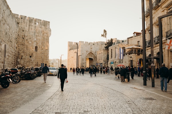 A bustling street scene featuring ancient stone walls and a group of people walking along a cobblestone path. Various motorcycles are parked on the side, and a prominent archway leads into the distance. The architecture is historical, with flags and outdoor seating visible along the street.