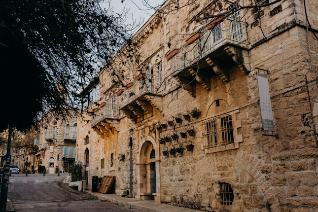 A narrow street lined with historic stone buildings featuring arched windows and balconies. The warm hues of the stone create a rustic and inviting atmosphere, while potted plants add a touch of greenery. The street is quiet, with minimal foot traffic and a parked car visible in the background.