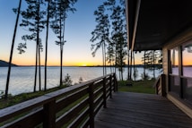 A serene lakeside view at sunset with tall, slender trees silhouetted against the colorful sky. A wooden deck extends from a house, providing an ideal spot to enjoy the tranquil scenery. The calm water reflects the colors of the sky, which transitions from deep blue to warm orange hues near the horizon.