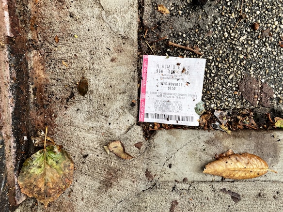 A lottery ticket is lying on a textured concrete surface beside fallen leaves, suggesting it is outdoors. The ground is a mix of dark, wet patches and dry, cracked cement with scattered debris around the ticket.