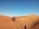 A caravan of camels guided by people traverses across expansive sand dunes under a clear blue sky. The golden hue of the desert landscape is highlighted by the shadow of a solitary individual on the right.
