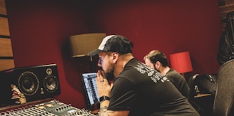 Two people are working in a music studio, with one person focusing intently on a computer screen and the other sitting beside him. The room features audio equipment, including large speakers and a control panel. The lighting is warm, and there is a red lamp providing ambient light.