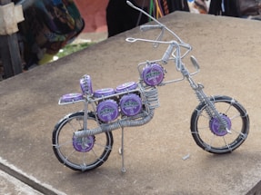 A miniature bicycle sculpture made from wire and decorated with purple bottle caps. The intricate design includes details like handlebars, seat, and wheels. The bottle caps feature the logo of a drink brand, adding a colorful contrast to the metallic wires.