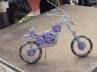 A miniature bicycle sculpture made from wire and decorated with purple bottle caps. The intricate design includes details like handlebars, seat, and wheels. The bottle caps feature the logo of a drink brand, adding a colorful contrast to the metallic wires.