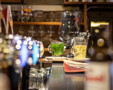 A warm and inviting scene inside a bar or caf&eacute;, with a focus on two steaming cups of herbal tea on the counter. The background features shelves lined with glasses and bottles, adding to the cozy atmosphere.