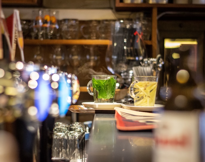 A warm and inviting scene inside a bar or café, with a focus on two steaming cups of herbal tea on the counter. The background features shelves lined with glasses and bottles, adding to the cozy atmosphere.