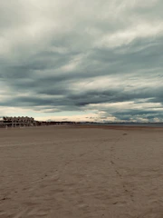 A vast sandy beach stretches out toward the horizon under a dramatic sky filled with thick, layered clouds. In the distance, a line of buildings and structures are visible, possibly hotels or apartments, suggesting a coastal urban setting. The beach area appears deserted, evoking a sense of solitude and calm.