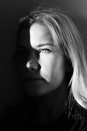 A black and white portrait of a person, partially illuminated by a light source creating dramatic shadows across their face. The lighting emphasizes one eye and part of the hair, while the rest of the face fades into shadow. The expression appears thoughtful or contemplative.