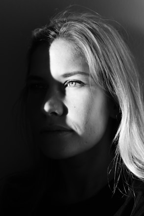 A black and white portrait of a person, partially illuminated by a light source creating dramatic shadows across their face. The lighting emphasizes one eye and part of the hair, while the rest of the face fades into shadow. The expression appears thoughtful or contemplative.