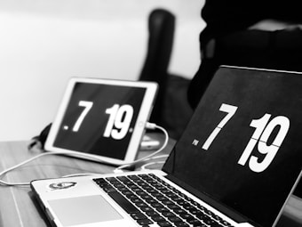 A laptop and a tablet displaying the time 7:19 with large white numbers on black screens are placed on a wooden table. Both devices are connected with a cable. The image is in black and white, giving it a modern and minimalist look.