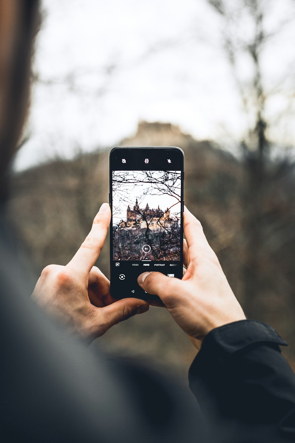 selective focus photo of phone hold by woman displaying bare trees and brown building in distant