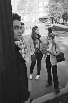 A group of young adults is standing together outdoors in an urban park area. The scene is in black and white, with several people in casual clothing. One person is holding a camera, another is using a smartphone, and two others are engaging in conversation. Trees and buildings are visible in the background, and scattered people can be seen in the park area.