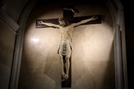 A dimly lit room features a detailed sculpture of a crucifix, highlighted by focused lighting. The figure is depicted with outstretched arms and a serene expression, surrounded by shadows that accentuate its features.