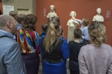 A group of people stands in front of several classical sculptures displayed in a museum. A woman gestures towards one of the busts while others observe closely. The setting has an educational atmosphere, and the sculptures are positioned against a red background.