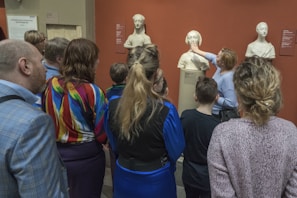 A group of people stands in front of several classical sculptures displayed in a museum. A woman gestures towards one of the busts while others observe closely. The setting has an educational atmosphere, and the sculptures are positioned against a red background.