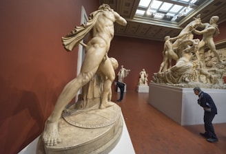 A gallery displaying classical sculptures with intricate details, depicting dynamic human figures in various poses. The room has a rich red color with a high ceiling and skylights. Visitors are observing the art pieces closely, suggesting an appreciation for the artistic craftsmanship.