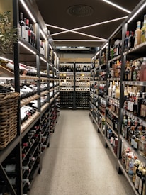 A well-organized liquor store aisle featuring a variety of wine and spirit bottles neatly arranged on metal shelves. The aisle has a modern, sleek design with bright lighting and a polished concrete floor. Baskets hold additional bottles on the left side, and signs above indicate regional categories such as Nordamerika and Europa.