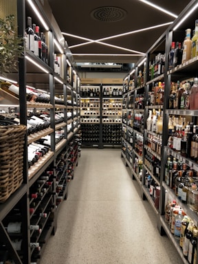 A well-organized liquor store aisle featuring a variety of wine and spirit bottles neatly arranged on metal shelves. The aisle has a modern, sleek design with bright lighting and a polished concrete floor. Baskets hold additional bottles on the left side, and signs above indicate regional categories such as Nordamerika and Europa.