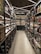 A well-organized liquor store aisle featuring a variety of wine and spirit bottles neatly arranged on metal shelves. The aisle has a modern, sleek design with bright lighting and a polished concrete floor. Baskets hold additional bottles on the left side, and signs above indicate regional categories such as Nordamerika and Europa.
