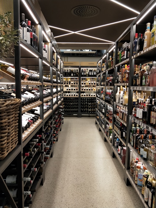 A well-organized liquor store aisle featuring a variety of wine and spirit bottles neatly arranged on metal shelves. The aisle has a modern, sleek design with bright lighting and a polished concrete floor. Baskets hold additional bottles on the left side, and signs above indicate regional categories such as Nordamerika and Europa.