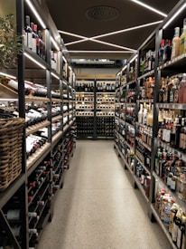 A well-organized liquor store aisle featuring a variety of wine and spirit bottles neatly arranged on metal shelves. The aisle has a modern, sleek design with bright lighting and a polished concrete floor. Baskets hold additional bottles on the left side, and signs above indicate regional categories such as Nordamerika and Europa.