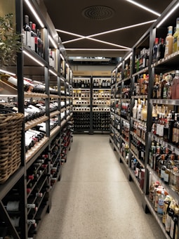 A well-organized liquor store aisle featuring a variety of wine and spirit bottles neatly arranged on metal shelves. The aisle has a modern, sleek design with bright lighting and a polished concrete floor. Baskets hold additional bottles on the left side, and signs above indicate regional categories such as Nordamerika and Europa.