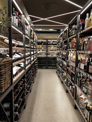 A well-organized liquor store aisle featuring a variety of wine and spirit bottles neatly arranged on metal shelves. The aisle has a modern, sleek design with bright lighting and a polished concrete floor. Baskets hold additional bottles on the left side, and signs above indicate regional categories such as Nordamerika and Europa.