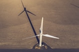 An aerial view of a large, white wind turbine standing on an expansive brown field. The shadow of the turbine extends across the textured surface of the land, creating a striking geometric pattern. The blades of the turbine are sharply defined and contrast with the earthen tones of the field. A small structure sits near the base of the turbine.