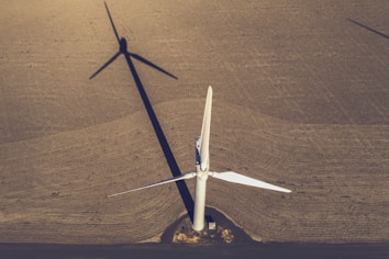 An aerial view of a large, white wind turbine standing on an expansive brown field. The shadow of the turbine extends across the textured surface of the land, creating a striking geometric pattern. The blades of the turbine are sharply defined and contrast with the earthen tones of the field. A small structure sits near the base of the turbine.