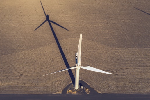 An aerial view of a large, white wind turbine standing on an expansive brown field. The shadow of the turbine extends across the textured surface of the land, creating a striking geometric pattern. The blades of the turbine are sharply defined and contrast with the earthen tones of the field. A small structure sits near the base of the turbine.