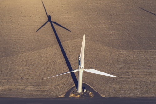 An aerial view of a large, white wind turbine standing on an expansive brown field. The shadow of the turbine extends across the textured surface of the land, creating a striking geometric pattern. The blades of the turbine are sharply defined and contrast with the earthen tones of the field. A small structure sits near the base of the turbine.