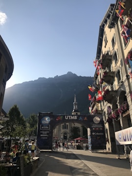 A picturesque street scene with a prominent building displaying various flags on the right. In the background, a mountain range is visible against a clear sky. In the center, there is an archway with 'UTMB' and logos, indicating an event location. People are walking around, with some seated at an outdoor café on the left.