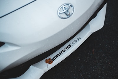 A close-up shot of the front of a white Toyota car, which prominently features the Toyota emblem. The car surface appears to have water droplets, suggesting it is wet from rain. A single brown fallen leaf rests on the car's bumper, which displays text reading 'INTROSPECTIVE DESIGN'. The background is a dark, wet surface, possibly asphalt.
