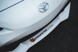 A close-up shot of the front of a white Toyota car, which prominently features the Toyota emblem. The car surface appears to have water droplets, suggesting it is wet from rain. A single brown fallen leaf rests on the car's bumper, which displays text reading 'INTROSPECTIVE DESIGN'. The background is a dark, wet surface, possibly asphalt.