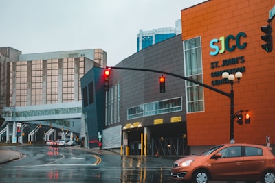An urban street scene under overcast skies featuring a modern building labeled SJCC, with St. John's Convention Centre in bold letters on an orange and gray facade. A wet street reflects the red light of a traffic signal, with a small compact car halted at the intersection. A skybridge connects two buildings, with a high-rise structure in the background.