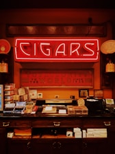 A vintage-style tobacco shop interior featuring a bright neon sign with the word 'Cigars' prominently displayed. Below it, a counter is filled with various cigar boxes and accessories. The walls are decorated with framed certificates and decorative signs, and there's an old-fashioned telephone and other shop items present.