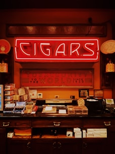 A vintage-style tobacco shop interior featuring a bright neon sign with the word 'Cigars' prominently displayed. Below it, a counter is filled with various cigar boxes and accessories. The walls are decorated with framed certificates and decorative signs, and there's an old-fashioned telephone and other shop items present.
