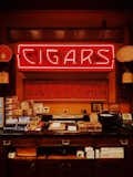 A vintage-style tobacco shop interior featuring a bright neon sign with the word 'Cigars' prominently displayed. Below it, a counter is filled with various cigar boxes and accessories. The walls are decorated with framed certificates and decorative signs, and there's an old-fashioned telephone and other shop items present.