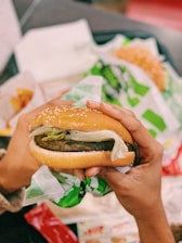 Hands holding a burger wrapped in paper, featuring a sesame seed bun, lettuce, and a patty. The background shows fast food wrappers and a hint of fries.