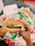 Hands holding a burger wrapped in paper, featuring a sesame seed bun, lettuce, and a patty. The background shows fast food wrappers and a hint of fries.