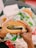 Hands holding a burger wrapped in paper, featuring a sesame seed bun, lettuce, and a patty. The background shows fast food wrappers and a hint of fries.