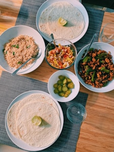 A dining table set with various dishes including flour tortillas with lime slices on top, a bowl of rice garnished with parsley, a colorful vegetable and corn salad, a dish of seasoned chicken with green bell peppers, and a small plate of sliced pickles. The setup includes a woven table mat and an empty glass.