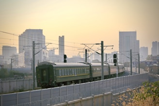 A train with green and beige coaches travels along a track, surrounded by modern city buildings under a hazy sky. Overhead electrical wires and signals are visible along the railway line.