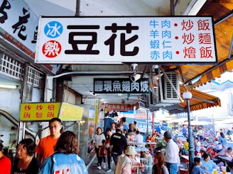 A bustling street market scene with a variety of signs displaying Chinese characters advertising food items. Numerous people are gathered, some standing and others seated at tables, engaging in conversations or enjoying meals. The environment is lively and colorful, with shades of red, blue, and other bright colors visible. The architecture suggests an outdoor market with awnings and signs hanging from the ceiling.