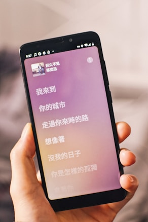 A hand is holding a smartphone displaying song lyrics in Chinese. The phone screen shows a music app with a pink and purple gradient background. The status icons like battery and WiFi are visible at the top.