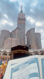 A blurred image of a large, iconic clock tower in the background with a clear view of the Kaaba in the foreground. In the immediate foreground, an open page of the Quran is visible. A group of people dressed in brightly colored clothing stands to the side.