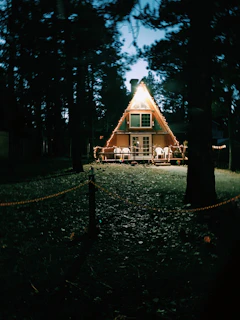 An A-frame cabin is adorned with string lights, creating a cozy glow. The surrounding environment is dark, filled with tall trees, and there is a wooden fence with a rope enclosing the area around the cabin.
